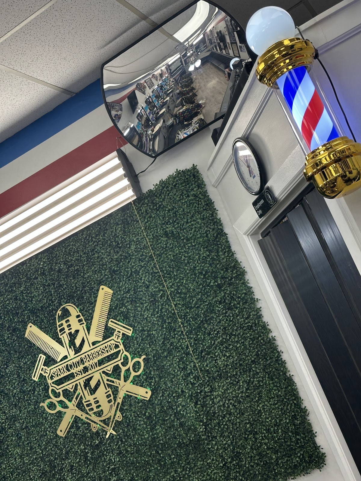 Gold Spark Cutz Barbershop Est. 2017 crest mounted on the greenery wall next to a lit barber pole